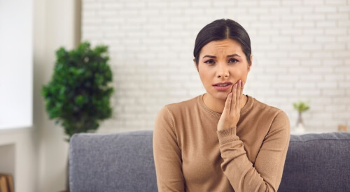 Wincing woman sitting on couch and holding her cheek in pain