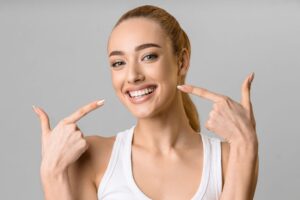 Woman with blond ponytail in white tank top pointing to her smile with 2 fingers